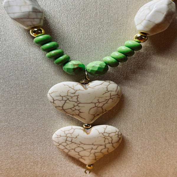A handmade beaded necklace featuring two large, vertically stacked heart-shaped beads with a white and brown crackle pattern, connected by a strand of green faceted beads and gold-toned spacers, displayed on a neutral beige textured fabric background.