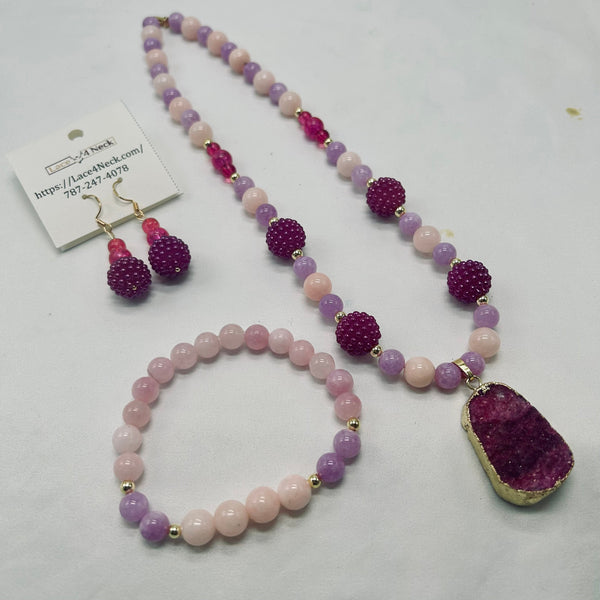 A jewelry set featuring a beaded necklace, matching beaded bracelet, and matching dangling earrings. The set includes light pink and lavender purple beads, textured purple crystal-like beads, and a large faceted purple geode pendant with a gold bezel.