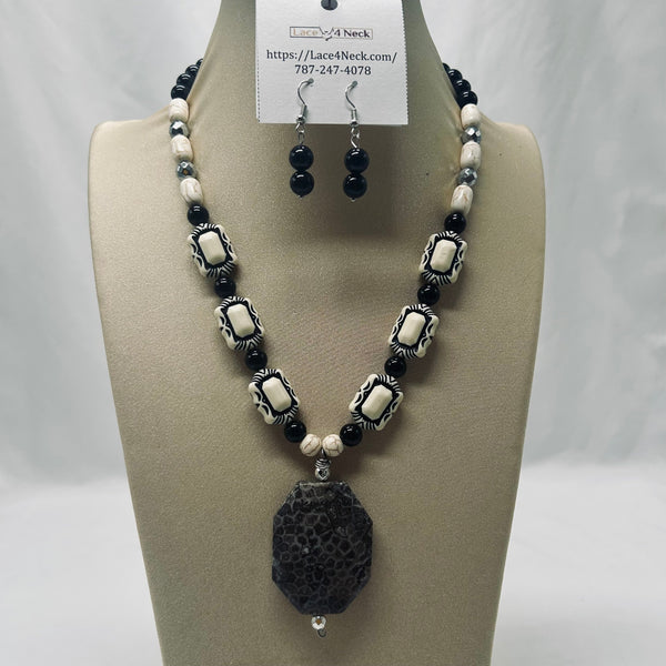 A beaded necklace and matching drop earrings displayed on a beige jewelry bust. The necklace features black beads, white beads, and silver-toned rectangular spacer beads with a large textured black oval pendant.