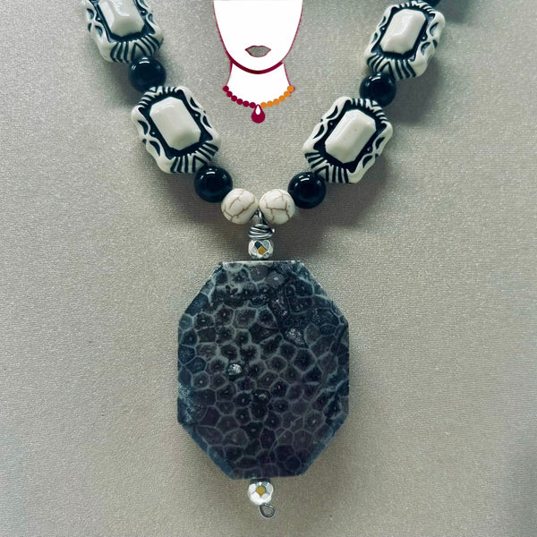 A beaded necklace featuring a large, textured dark gray agate pendant, white and black patterned rectangular beads, and small black round beads, displayed on a neutral gray background.