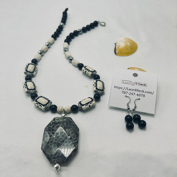 A beaded necklace and matching drop earrings displayed on a white surface, with a small shell prop and a product tag. The necklace features a large faceted black stone pendant, black beads, and white beads with black decorative accents.