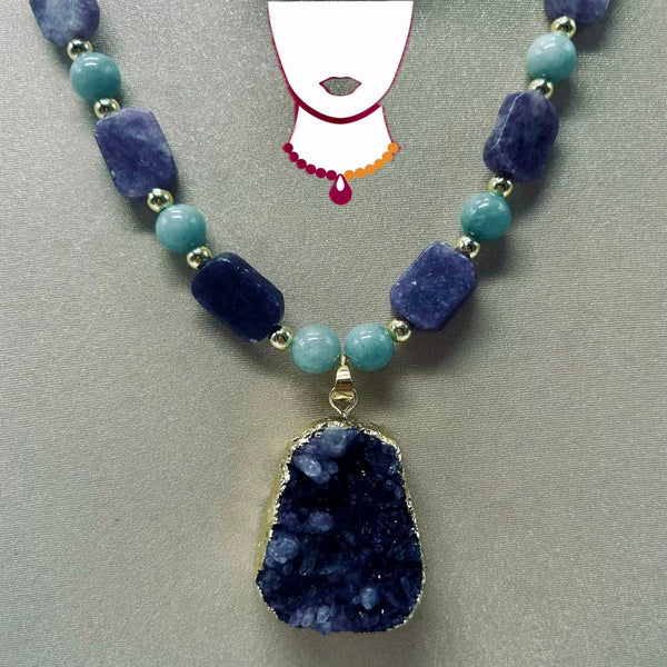 A beaded necklace featuring dark blue polished stone beads, light blue round beads, and a large dark blue geode pendant with a gold-tone border, displayed on a neutral gray background.
