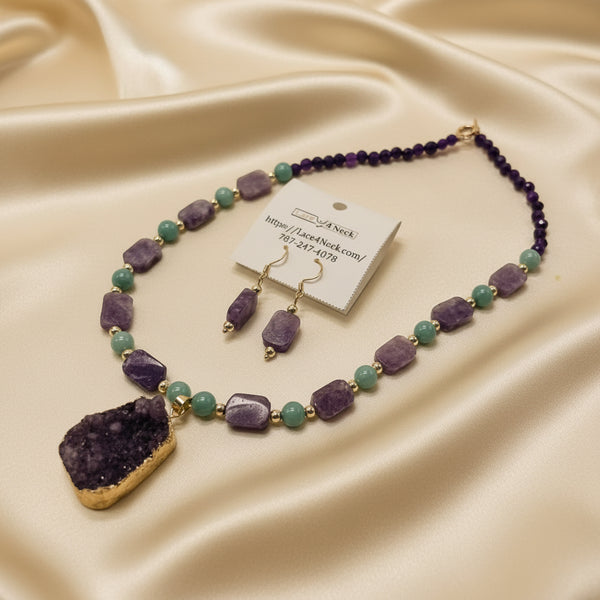 A beaded necklace and matching drop earrings displayed on a white surface. The jewelry features lavender-purple and light teal beads with gold-tone metal accents, including a large irregular purple geode pendant on the necklace and matching purple stone drops on the earrings.