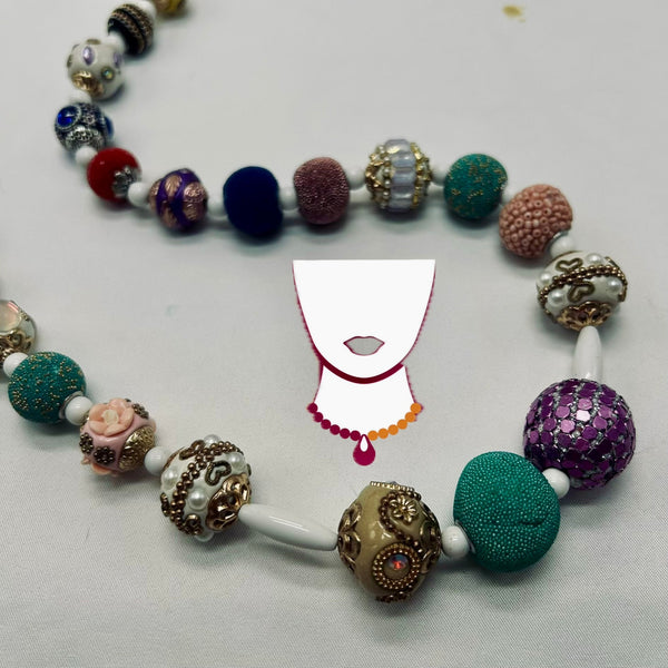 A handmade beaded necklace with a variety of colorful, textured, and ornate glass beads in green, purple, pink, blue, and gold, displayed on a light gray fabric background with a white graphic overlay of a necklace template.