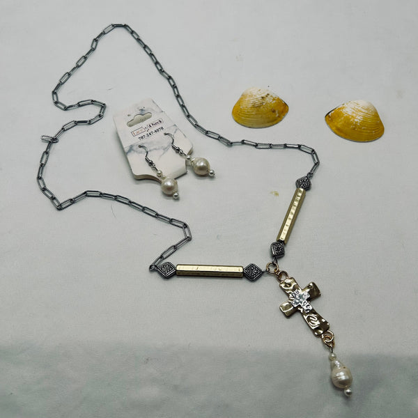 A gold and silver chain necklace with a cross pendant, bar links, and a dangling pearl, displayed alongside a matching pair of pearl drop earrings on a card. Two yellow seashells are placed behind the jewelry on a light gray surface.