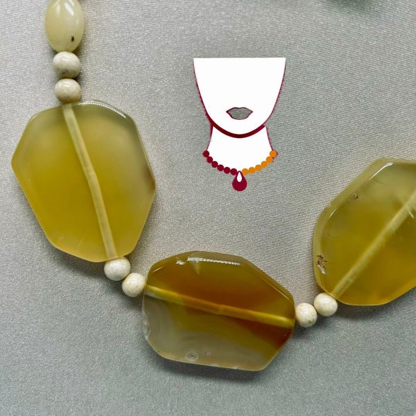 A close-up of a beaded necklace with large translucent yellow-green stone beads and small white spacer beads, laid on a gray textured surface. A small white graphic of a necklace is centered above the beads.