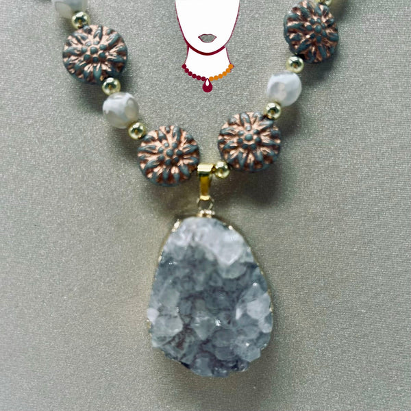 A beaded necklace featuring a large, teardrop-shaped gray geode pendant, accented with textured bronze flower beads, white pearls, and small gold spacer beads, displayed on a neutral gray background.