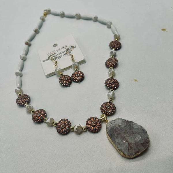 A beaded necklace and matching drop earrings displayed on a light gray surface. The necklace features white translucent beads, textured metallic rose-gold floral medallions, and a large natural druzy stone pendant.