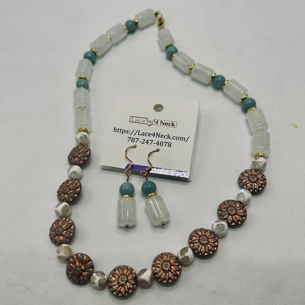 A beaded necklace and matching drop earrings displayed on a white surface with a product tag. The necklace features translucent white cylindrical beads, small teal-blue beads, and rose-gold-toned spacer accents, with larger rose-gold floral disc beads.