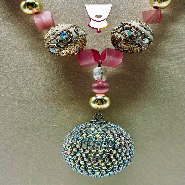 A close-up of a beaded necklace or ornament featuring ornate gold-toned filigree beads, translucent pink beads, clear faceted beads, and a large iridescent rhinestone-covered spherical charm hanging at the bottom, set against a plain taupe background.
