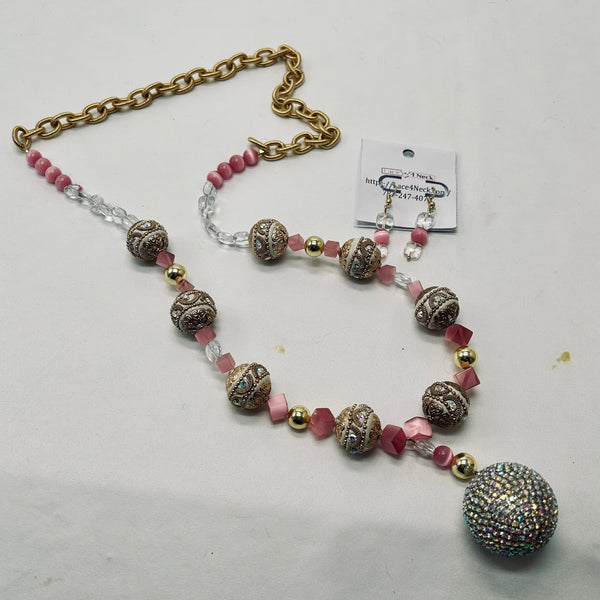 A beaded necklace and matching drop earrings laid out on a light off-white surface. The jewelry features pink faceted beads, clear crystal beads, textured metallic bronze and gold spacer beads, and a large iridescent rhinestone-covered ball at the end of the necklace.