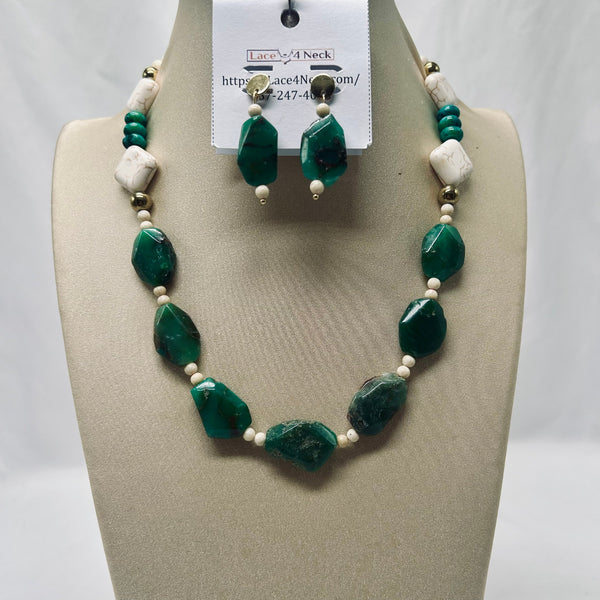 A beaded necklace and matching drop earrings displayed on a beige jewelry bust. The necklace features large faceted green stones, small white beads, and white textured cube beads, with gold-toned metal findings.
