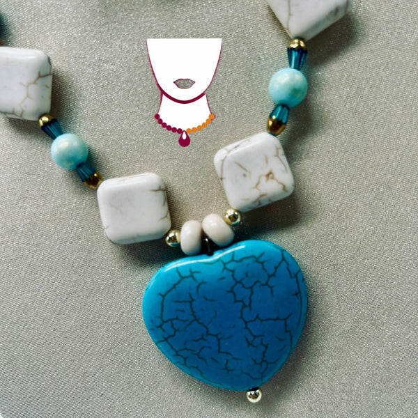 A beaded necklace featuring a large, vibrant blue heart-shaped pendant with a black crackle pattern, accented by white marbled square beads, light blue round beads, and small gold spacer beads, displayed on a textured gray background.