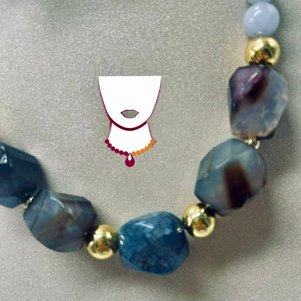 A beaded necklace with faceted blue and gray agate-like beads, polished gold spacer beads, and a pale lavender bead, displayed on a textured gray background with a small stylized necklace graphic.