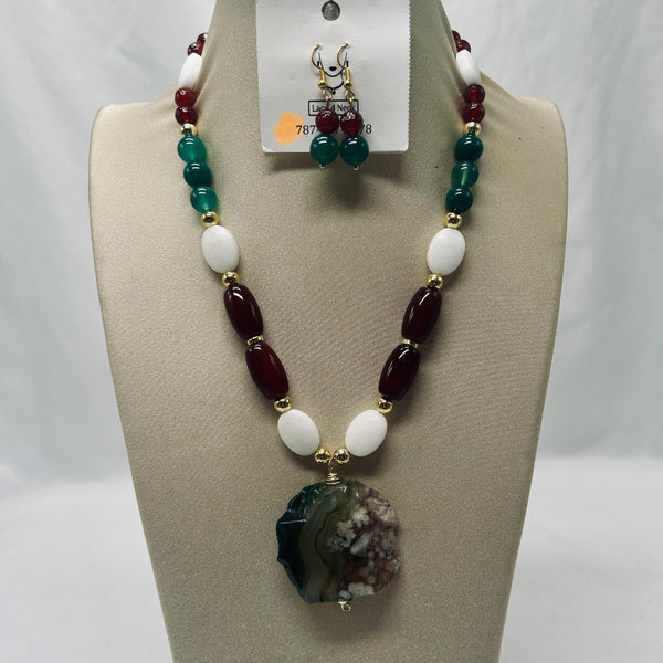 A beaded necklace displayed on a beige jewelry bust, with a matching pair of drop earrings hanging from a card above it. The necklace features dark red oval beads, white oval beads, green round beads, and a large natural agate geode pendant.