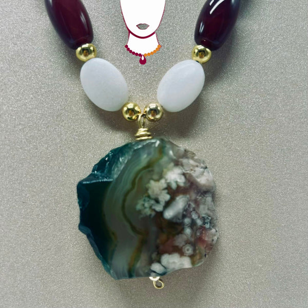 A beaded necklace featuring a large, raw agate geode pendant with green, white, and brown banding, accented by white oval beads, gold spacers, and dark red oval beads at the top.