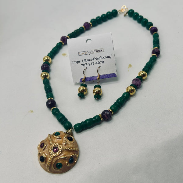 A beaded necklace and matching drop earrings displayed on a white surface. The necklace features dark green beads, purple beads, and gold spacer beads with a large round gold pendant accented by green and purple stones.