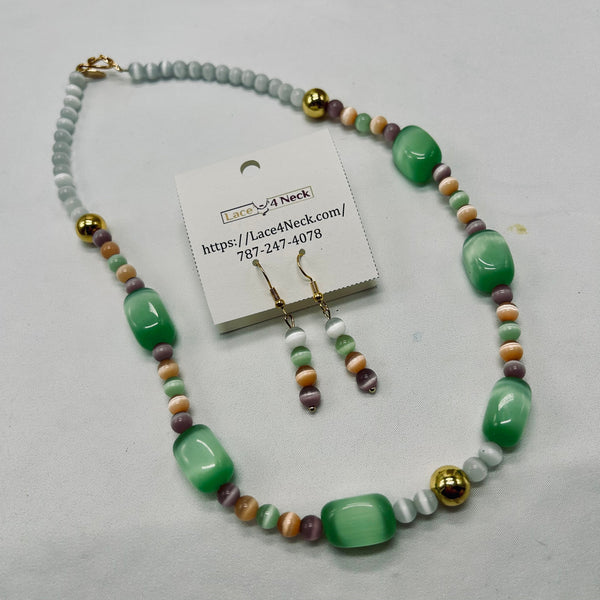 A beaded necklace and matching drop earrings displayed on a white card. The necklace features large green cabochon beads, small pale blue-gray beads, and multi-colored beads in peach, purple, and brown, accented with gold-tone spacer beads.