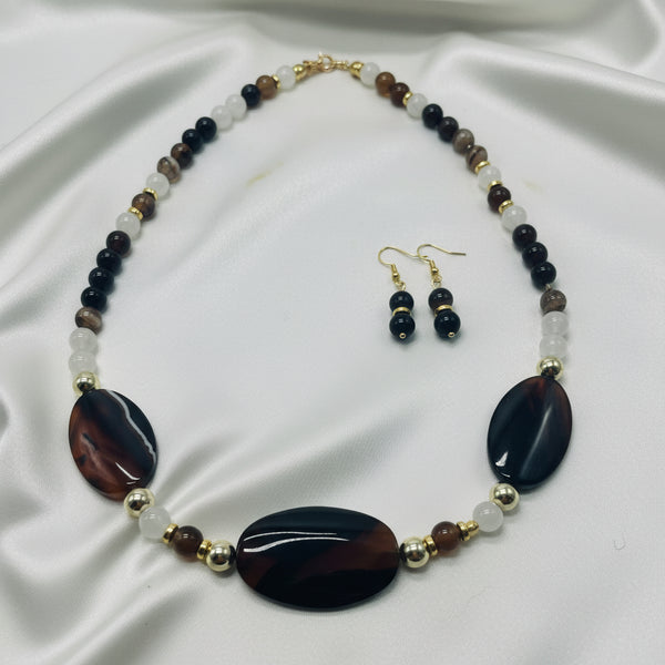 A beaded necklace and matching drop earrings displayed on a white surface. The necklace features two large oval dark brown and black marbled beads, alternating with small white beads, brown beads, and gold-tone spacer beads.