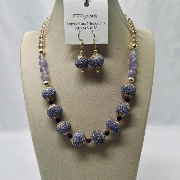 A beaded necklace and matching drop earrings displayed on a beige jewelry bust. The necklace features textured lavender-purple and gold beads, with a white paper tag attached that reads 'Lace4Neck' and includes a phone number.