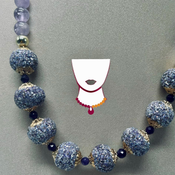 A beaded necklace with large textured blue-gray round beads, smaller dark blue beads, and gold-toned spacer details, displayed on a gray fabric background. A simple white necklace silhouette graphic is overlaid in the center.