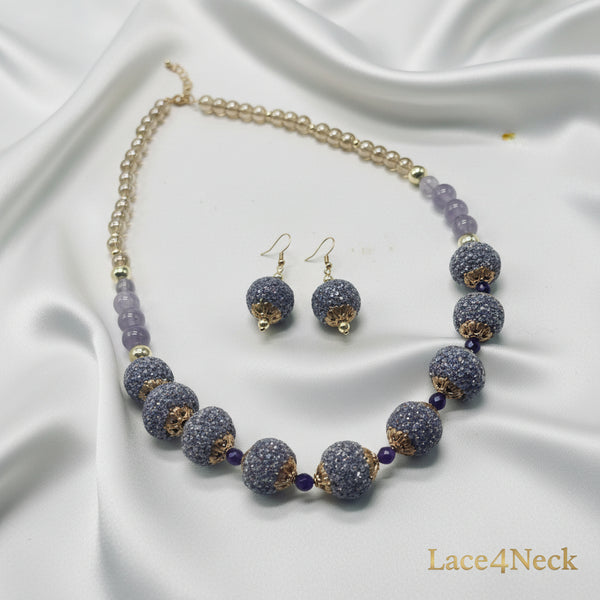 A beaded necklace and matching drop earrings displayed on a white surface with a product tag. The necklace features large, round, glittery lavender-purple beads with gold-toned filigree accents and small dark purple beads, alongside a chain of clear faceted beads and a gold-toned clasp.