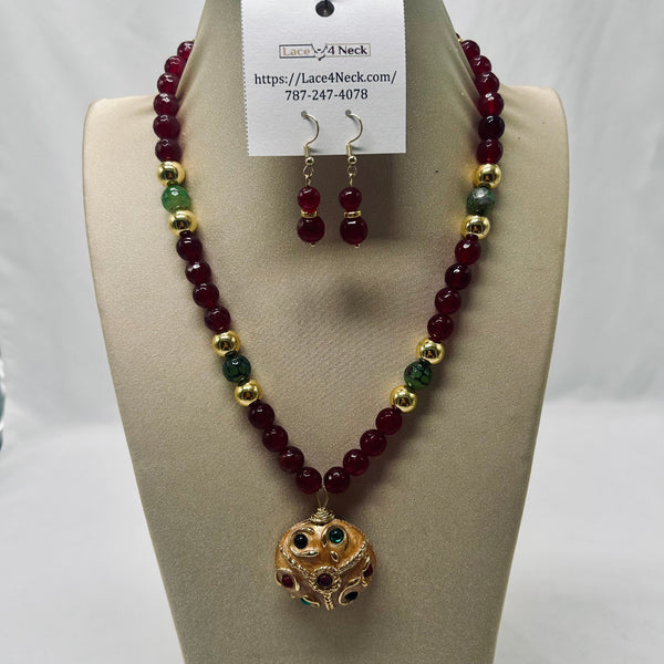 A beaded necklace and matching drop earrings displayed on a beige jewelry bust. The necklace features deep red beads with alternating gold and green beads, and a gold-toned owl pendant with red and green accents.