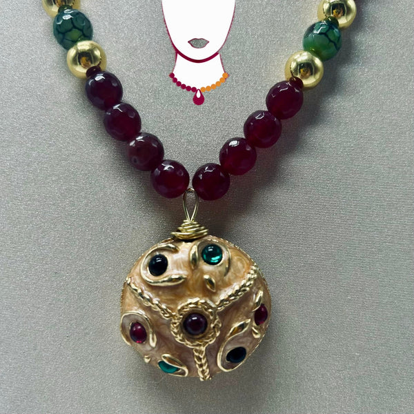 A beaded necklace with dark red faceted beads, green marbled beads, and gold-toned accents, featuring a large ornate gold pendant with red and green gemstone details, displayed on a gray textured background.