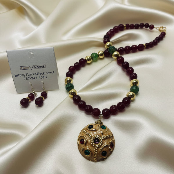 A beaded necklace and matching drop earrings displayed on a white surface, with a small branded card. The necklace features dark red beads, green beads, and gold-toned spacer beads, with a round ornate gold-toned pendant containing small red and green stones.