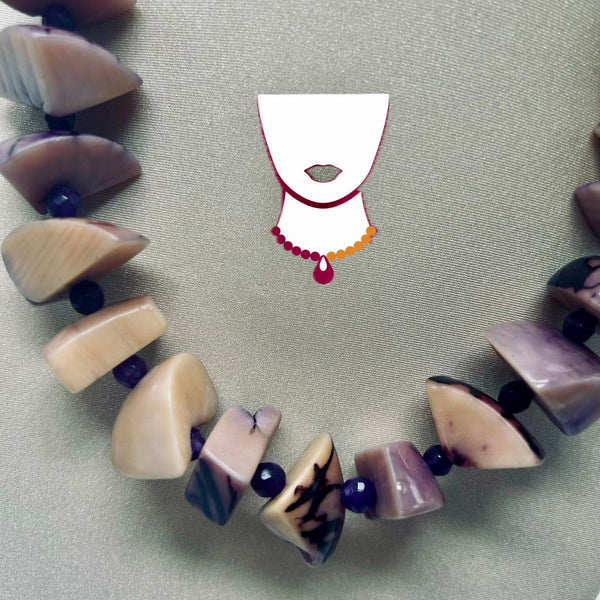A beaded necklace made of irregular polished shell or stone pieces in cream, beige, lavender, and gray tones, separated by small dark beads, laid on a light gray textured surface. A small white necklace-shaped graphic with red and orange trim appears near the center.