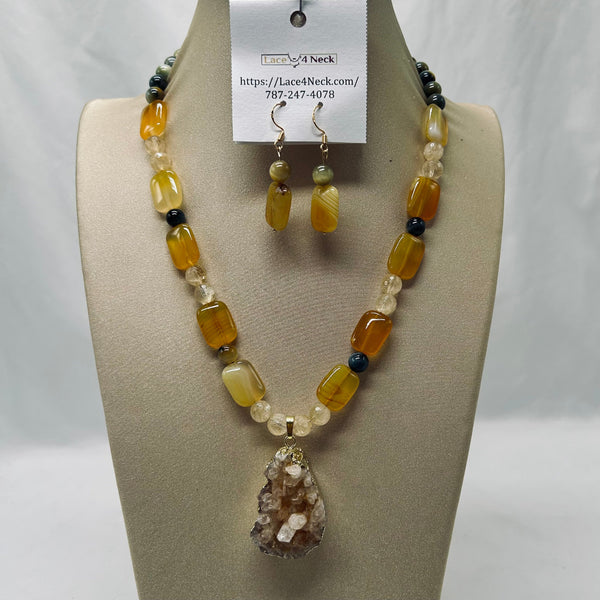 A beaded necklace and matching drop earrings displayed on a beige jewelry bust. The necklace features faceted amber-yellow, cream, and dark gray beads with a teardrop crystal pendant.