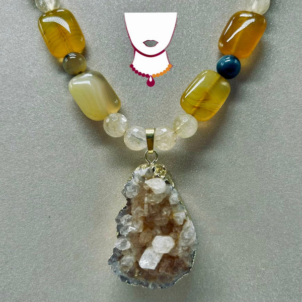A beaded necklace with a natural geode cluster pendant, featuring translucent yellow agate, cream quartz, a dark blue bead, and a white graphic of a necklace at the top center.