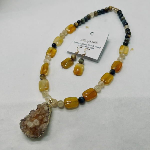 A beaded necklace with a large geode pendant and matching drop earrings, displayed on a white background with a product tag attached. The beads are primarily amber-yellow, cream, and dark brown, with a brown geode cluster pendant.