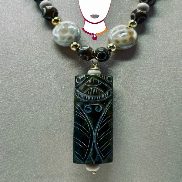A beaded necklace featuring a prominent rectangular carved pendant in a dark greenish-black stone with intricate etched patterns. The pendant is suspended from a strand of alternating polished agate beads in cream, brown, and black, accented with small gold spacer beads, all displayed on a neutral gray background.