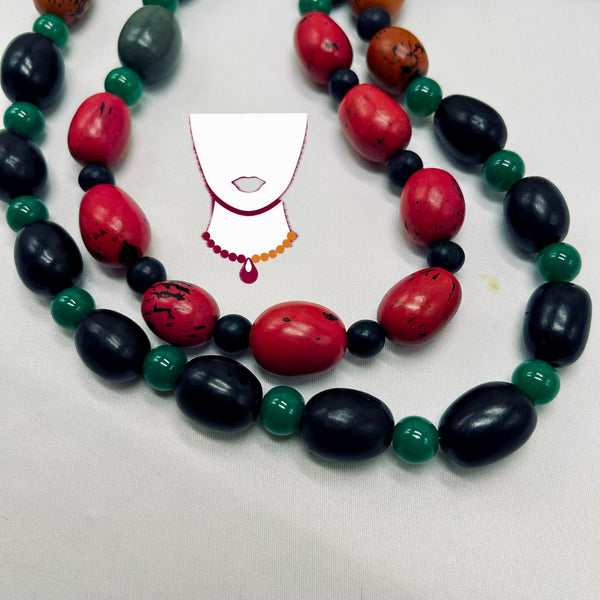A beaded necklace with large polished red and black oval beads, smaller green round beads, and a few brown speckled beads, arranged in a loose circle on a light gray background. A simple white necklace graphic with red lips and a dangling red teardrop sits near the center.