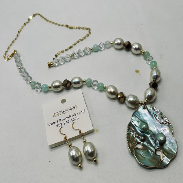 A necklace and matching drop earrings displayed on a light gray surface. The necklace has a gold-tone chain, clear faceted beads, pale mint-green beads, large silver-gray pearls, and a large irregular iridescent shell pendant.