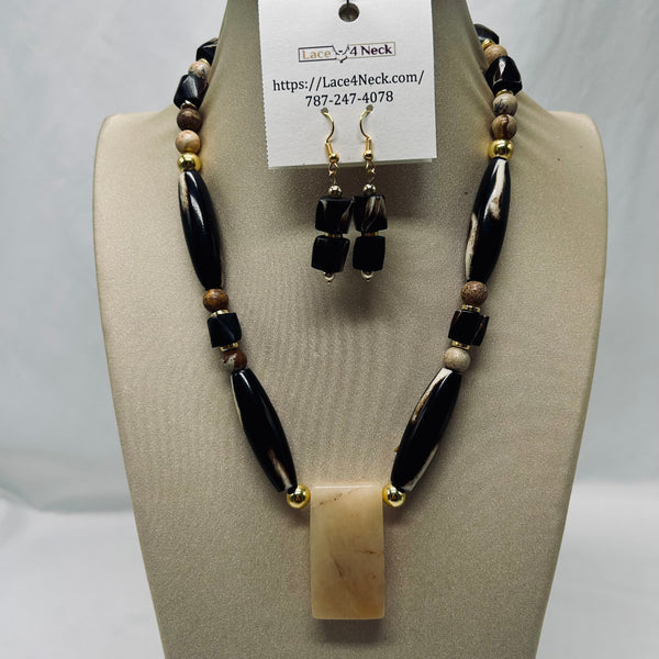 A beaded necklace and matching drop earrings displayed on a beige jewelry bust. The necklace features glossy black and white marbled beads, small brown and cream spacer beads, and a rectangular cream-colored pendant with gold-tone ends.