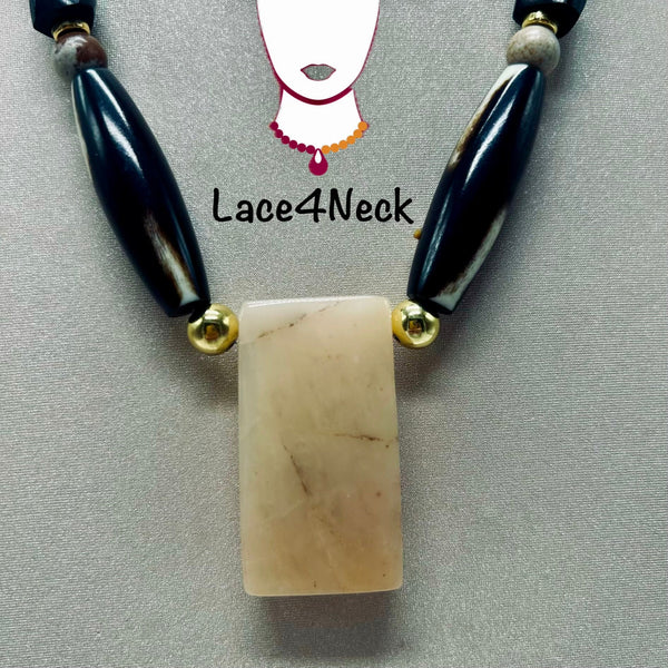A beaded necklace featuring a rectangular cream-colored stone pendant, flanked by glossy black and white beads with gold spacers, displayed on a gray textured background with a Lace4Neck logo overlay.
