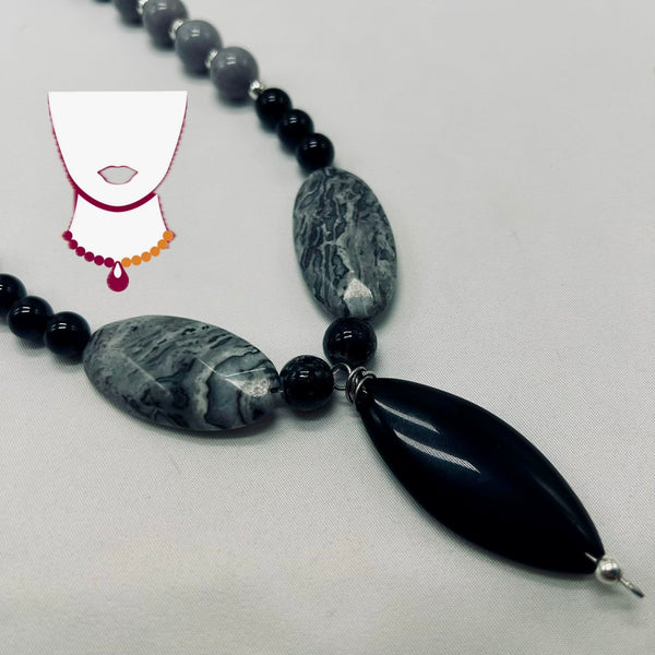 A beaded necklace featuring large polished gray marbled oval beads, smaller black round beads, and a large glossy black teardrop-shaped pendant, displayed on a light gray fabric background.