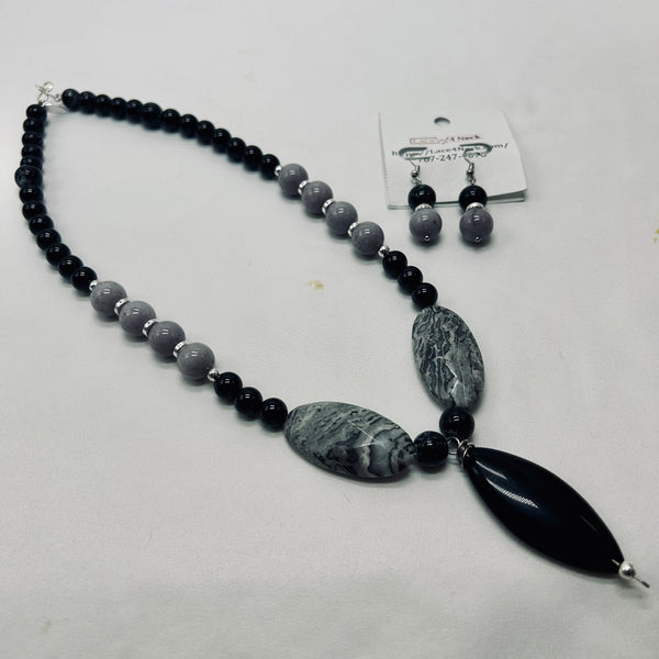A beaded necklace and matching drop earrings displayed on a light gray background. The necklace features black beads, gray beads, two oval gray stone pendants, and a large black teardrop stone pendant.