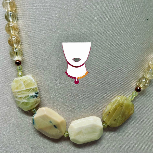 A beaded necklace displayed on a textured gray background, featuring large cream and olive-green faceted beads, clear crystal beads, and small metallic brown beads. A small stylized graphic of a white neck with red and orange trim is centered above the necklace.