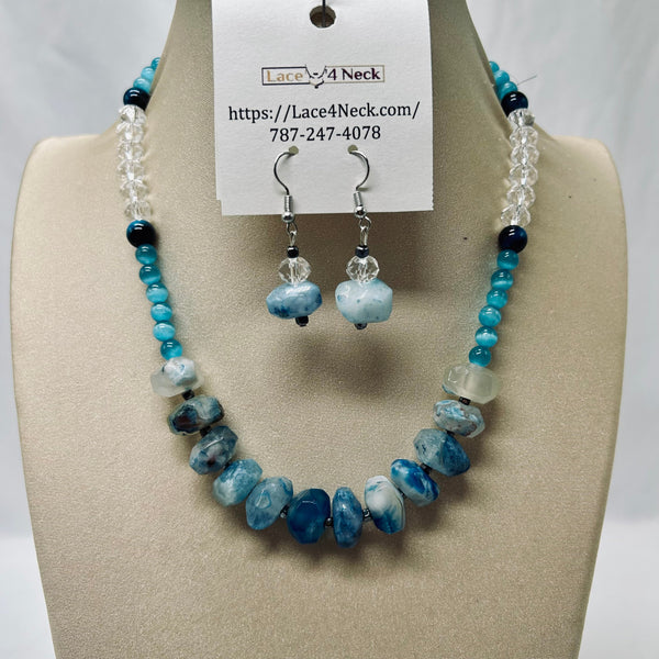 A beaded necklace and matching drop earrings displayed on a beige jewelry bust. The necklace features turquoise blue beads, marbled blue and white stones, clear faceted beads, and dark navy beads, with a white paper tag attached that reads Lace4Neck.