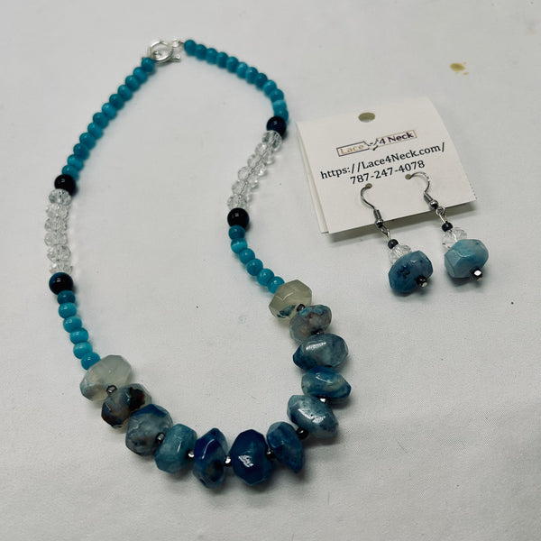 A beaded necklace and matching drop earrings displayed on a white surface. The necklace features turquoise blue beads, faceted blue and gray stones, clear crystal beads, and small black beads, with a silver clasp.