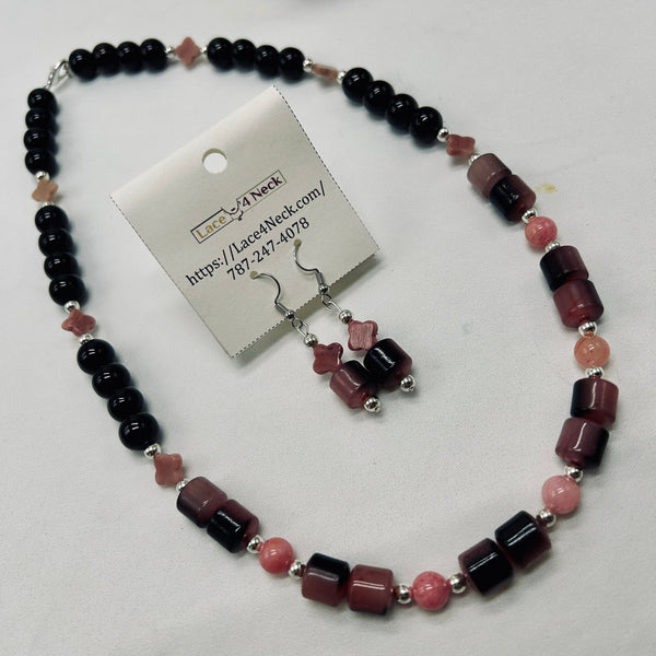 A beaded necklace and matching drop earrings displayed on a white surface, with a branded card attached to the necklace. The jewelry features glossy black round beads, faceted dark pink and burgundy rectangular beads, and small pink star-shaped beads separated by silver spacer rings.