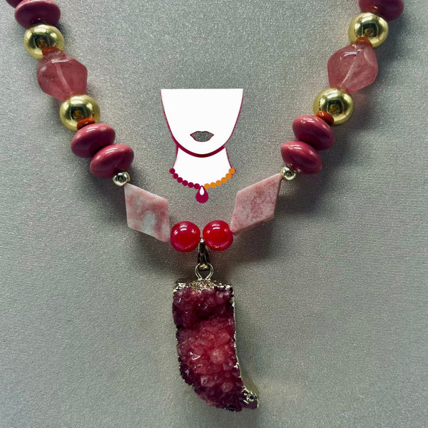 A beaded necklace featuring pink and red gemstone beads, polished gold spacer beads, two pink crystal chips, and a large pink geode cluster pendant, displayed on a gray background with a white necklace display form behind it.